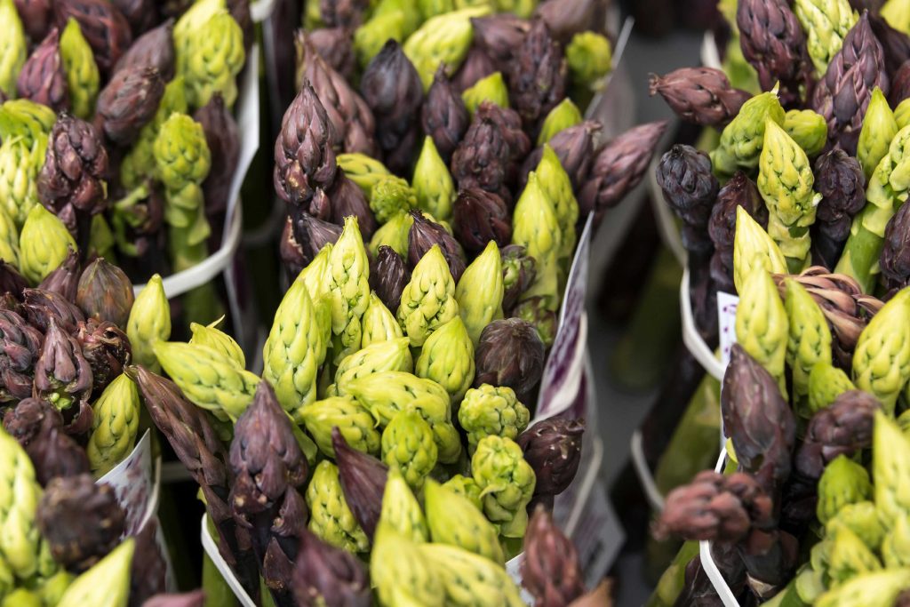Close-up of fresh green and purple asparagus spears bundled together, displayed at a farmers market or produce stand.