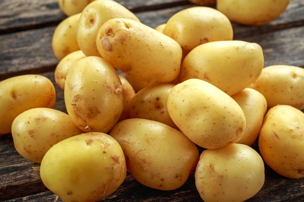 A collection of freshly harvested Jersey Royal potatoes with light golden skin and soil specks, arranged on a dark wooden surface.