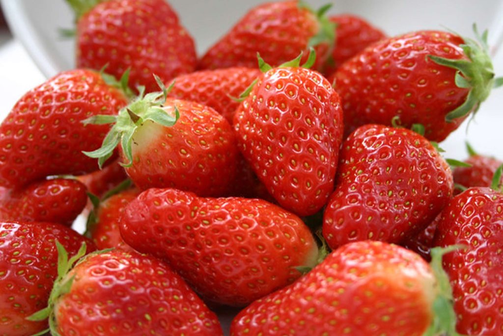 A close-up photograph of fresh, ripe red strawberries with green leafy tops arranged together on a white background.